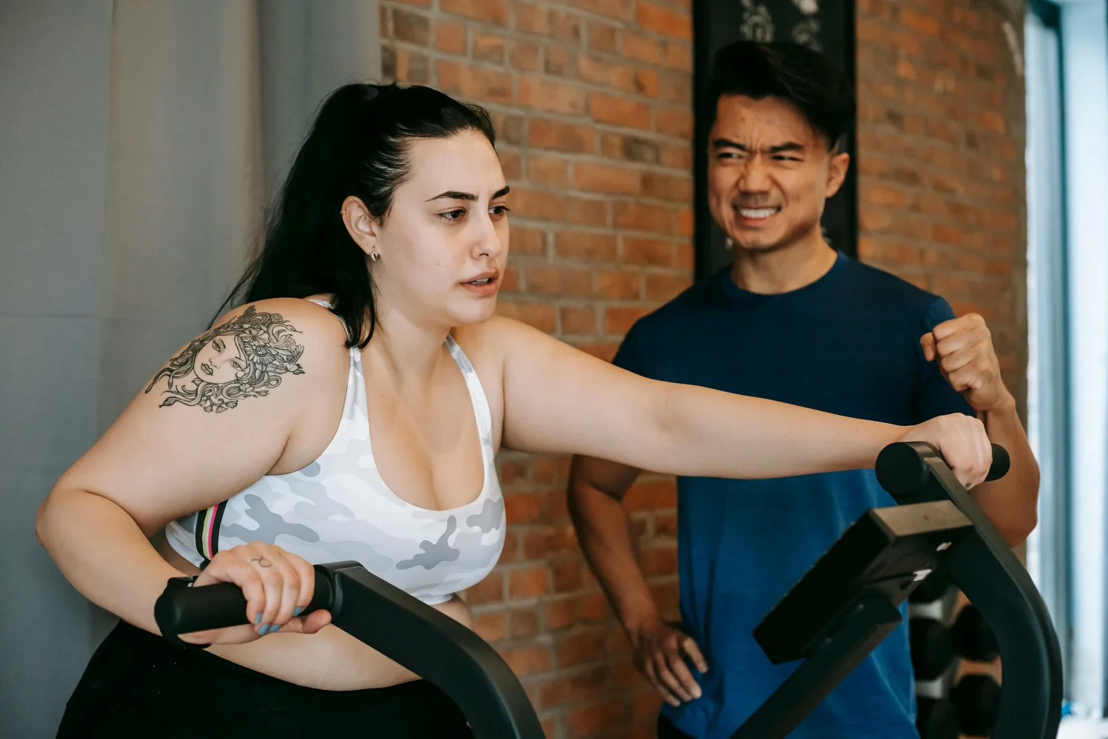 A woman on a cycling machine with a man standing beside her in a brick-walled gym