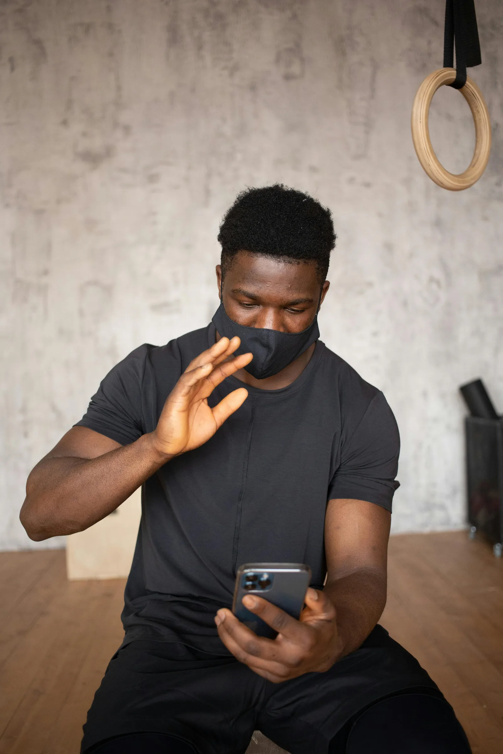 A man in a black t-shirt sitting on a gym floor looking at his phone