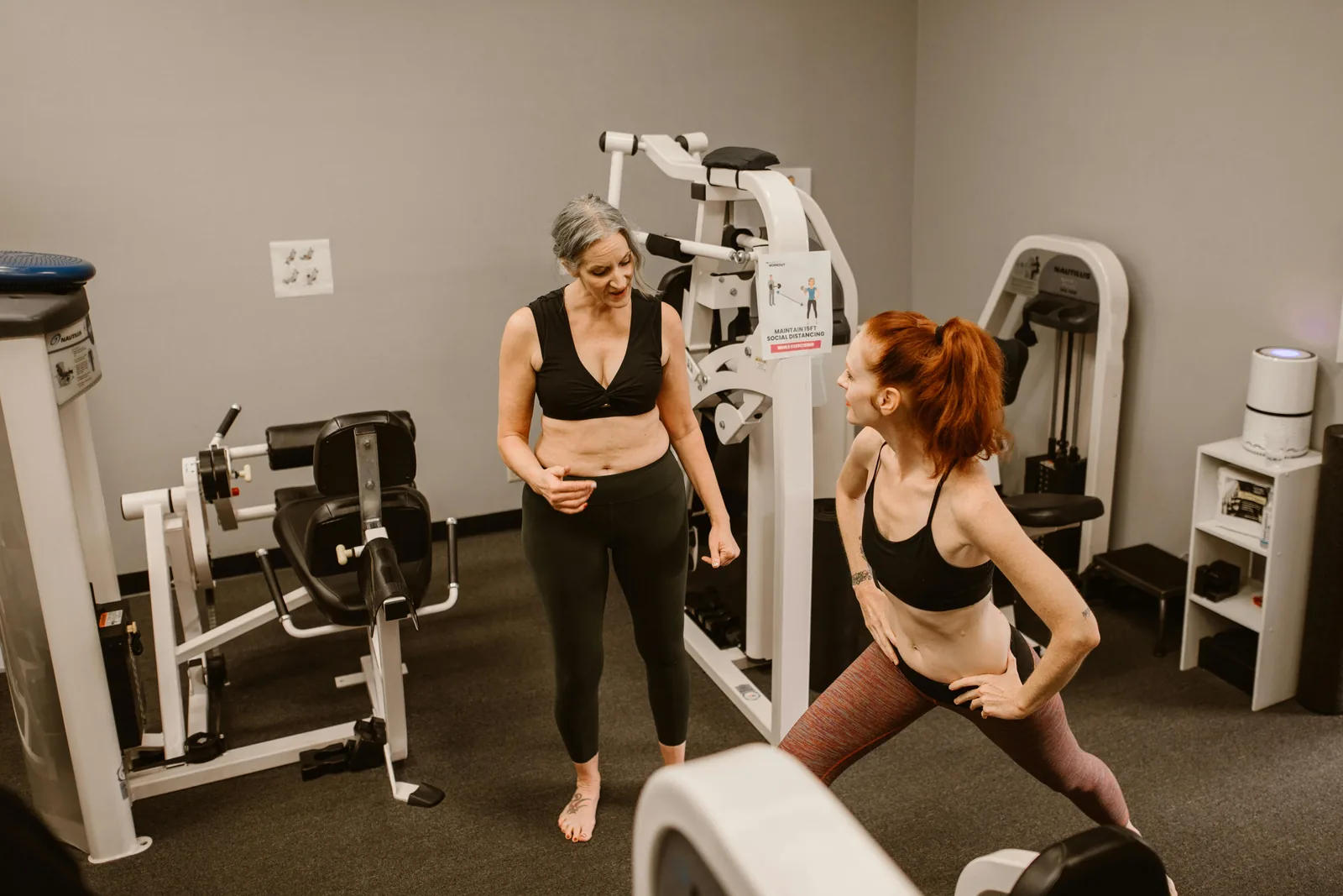 An older woman and a younger woman standing together between weight machines in a small gym